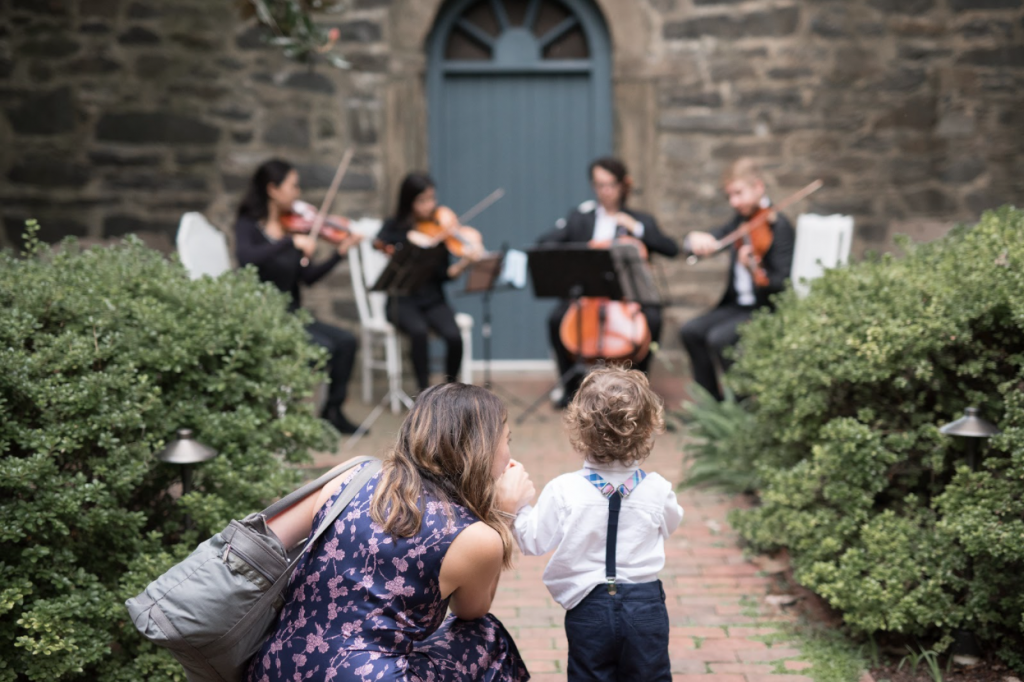 A wedding guest and her toddler son relax next to the wedding string quartet as they play a custom playlist during a wedding prelude.